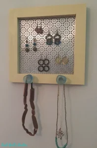 A picture framed that has been filled with decorative screening from which earrings hang. It also has two knobs along the bottom that hold necklaces.