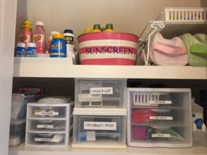 items on bathroom shelves, including sunscreen, hair bands, and medications, each contained in labeled bins and drawers.