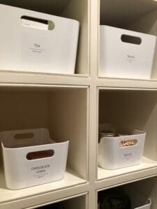White pantry bins holding tea, treats, chocolate chips, snacks, sitting on cubby shelves.